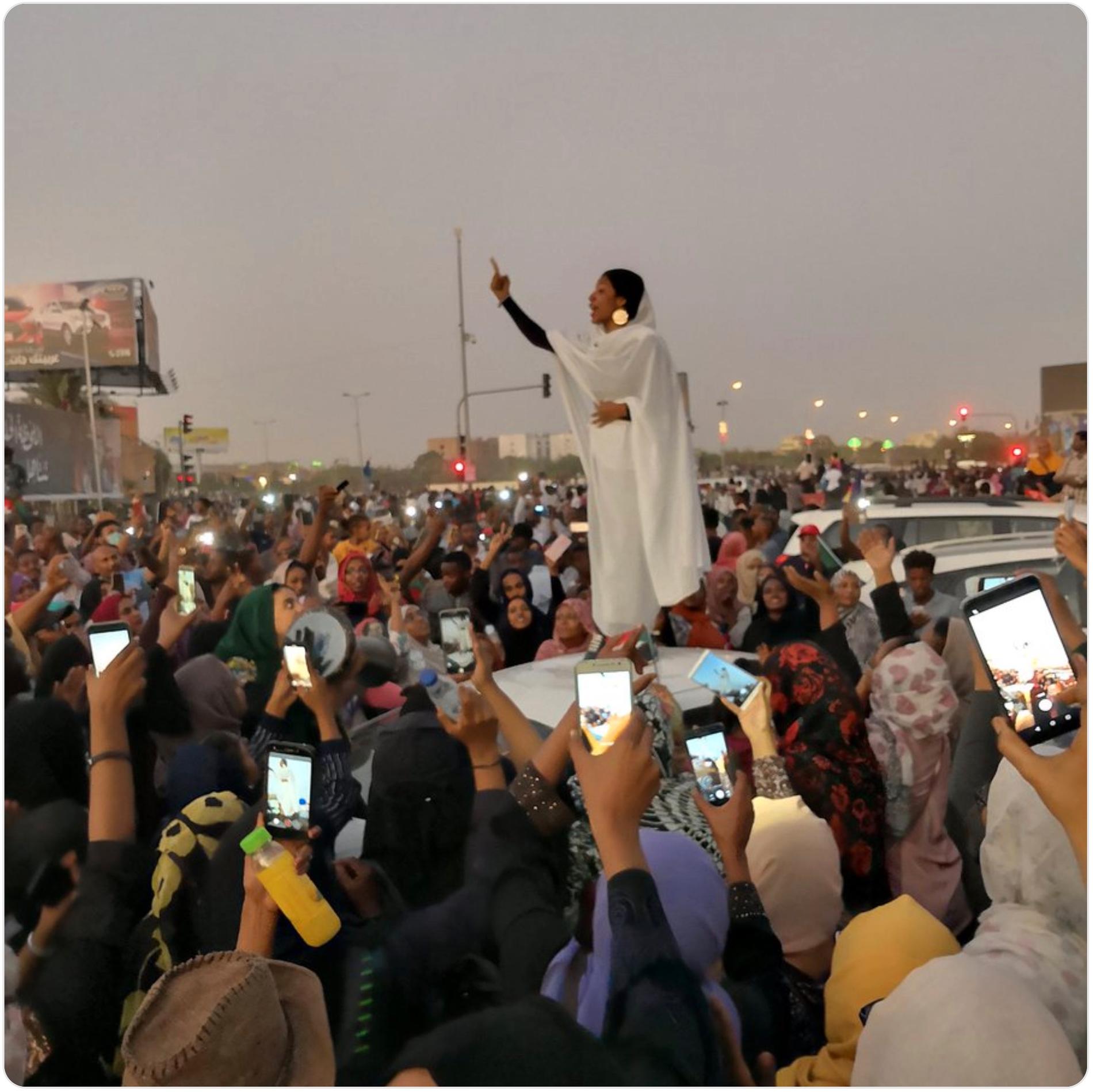 Den unga sudanesiska kvinnan Alaa Salah, 22, står på en bilmotorhuv i huvudstaden Khartoum. Hon har höjt ena armen och har ett finger i luften, och leder protestsången med hjälp av alla de människor som samlats kring henne.