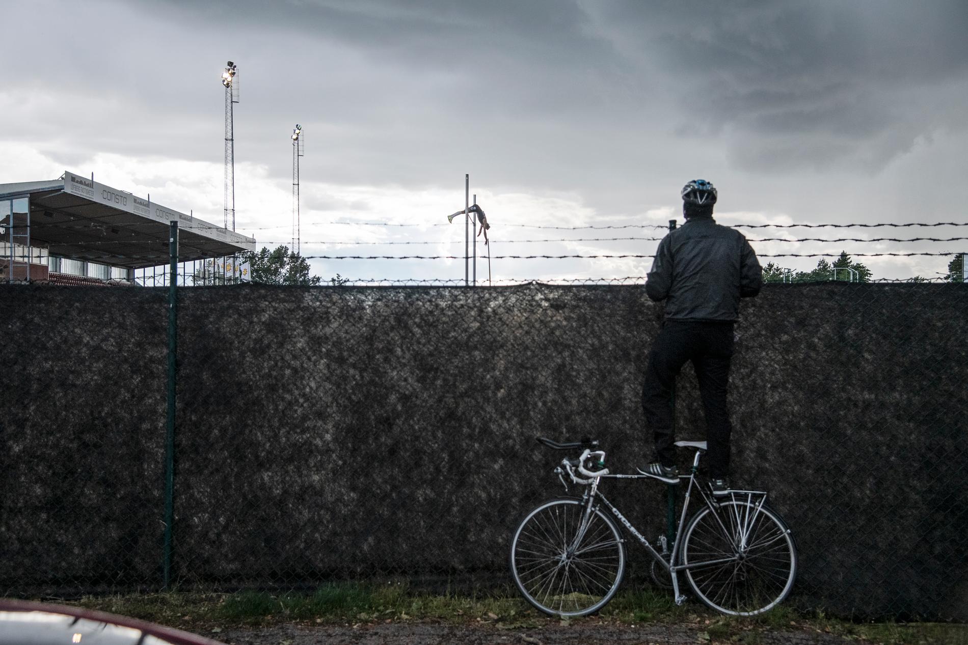 En man står på sin cykel för att se över staketet när Armand Duplantis vinner herrarnas stavhopp på 5,72 meter i friidrottstävlingen Karlstad GP den 8 juli 2020 på Tingvalla IP. På grund av corona-pandemin tillåts ingen publik på arenan.