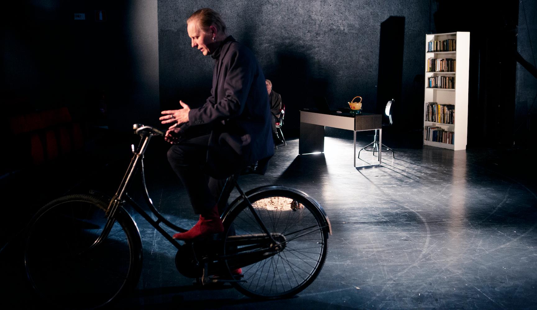 Andreas Boonstra på cykel och i bakgrunden skymtar Linda Källgren i moment:teaters ”Doggerland – att läsa Julia Blackburn”.
