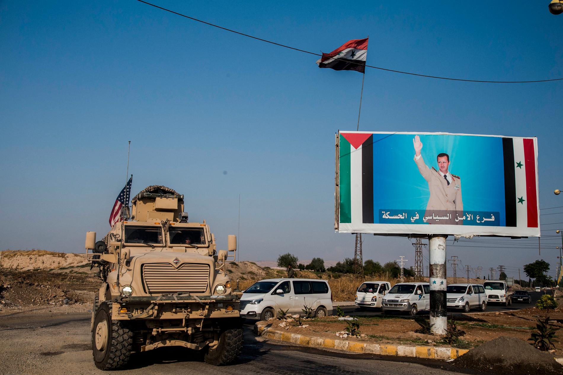 En amerikansk militärkonvoj i staden Qamishli i norra Syrien bredvid en bild av landets president Bashar al-Assad. Arkivbild.
