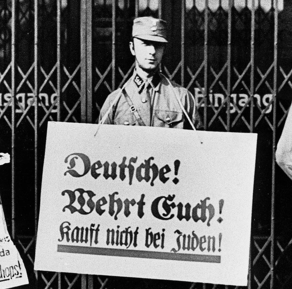 En nazist står utanför en judisk butik med plakat som uppmanar folk att inte handla hos judar. I dag är det 75 år sedan koncentrationslägret i Auschwitz befriades, vilket uppmärksammas med den internationella minnesdagen för förintelsens offer.