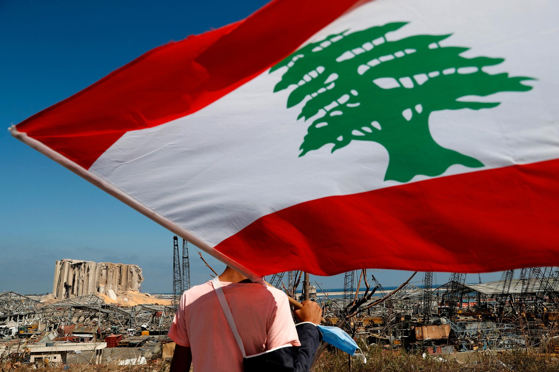 En man med den libanesiska flaggan tittar ut över förödelsen i hamnen efter tisdagens explosion i Beirut.