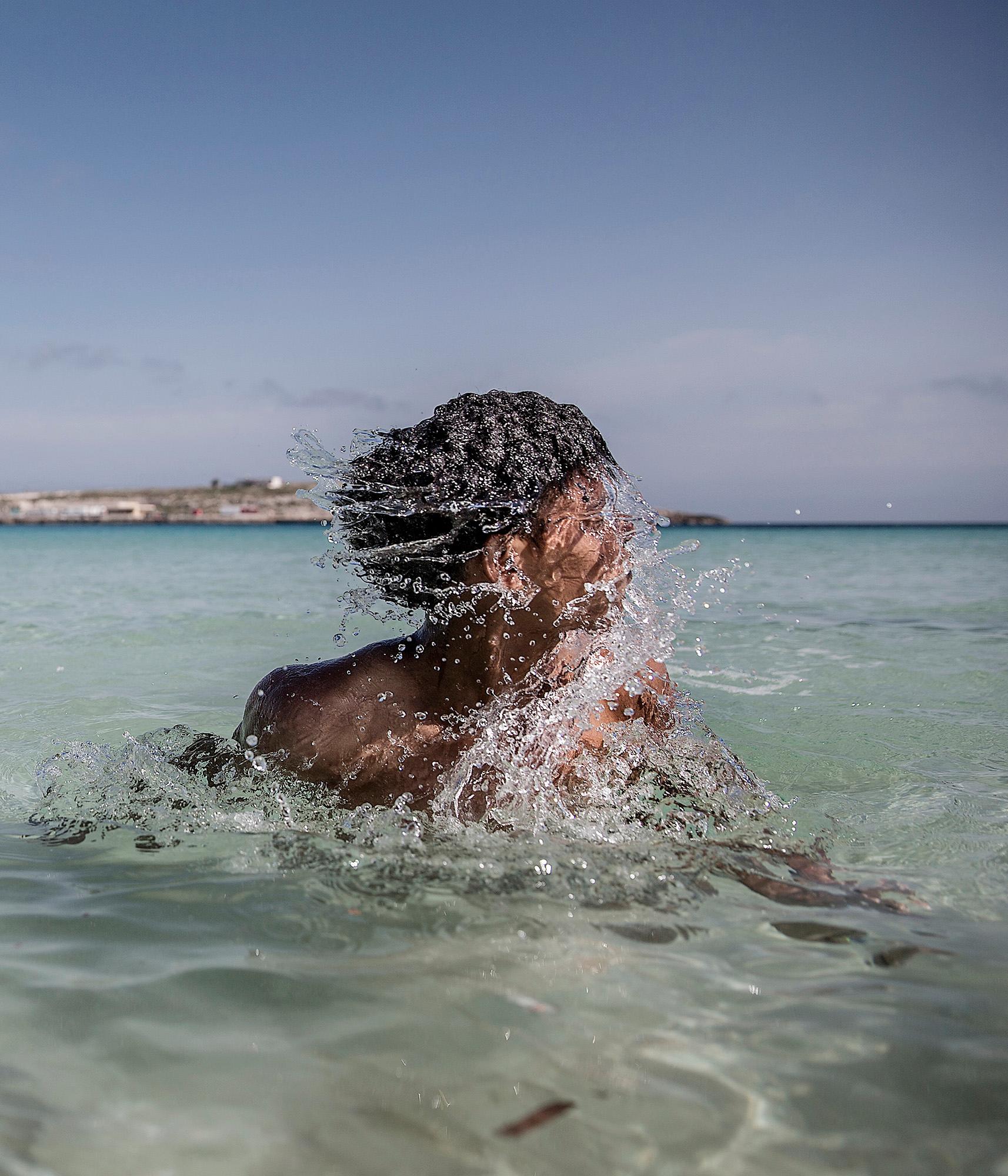Lampedusa, ön som ligger mitt i medelhavet och blivit en av de första anhalterna för båtflyktingar.  Robel, Nohom, Meron, Anbesa, Amanuel från Eritrea badar i det kristallblåa vattnet på en av lampedusas stränder. Under dagarna har de möjlighet att lämna det inhägnade lägret de bor i.