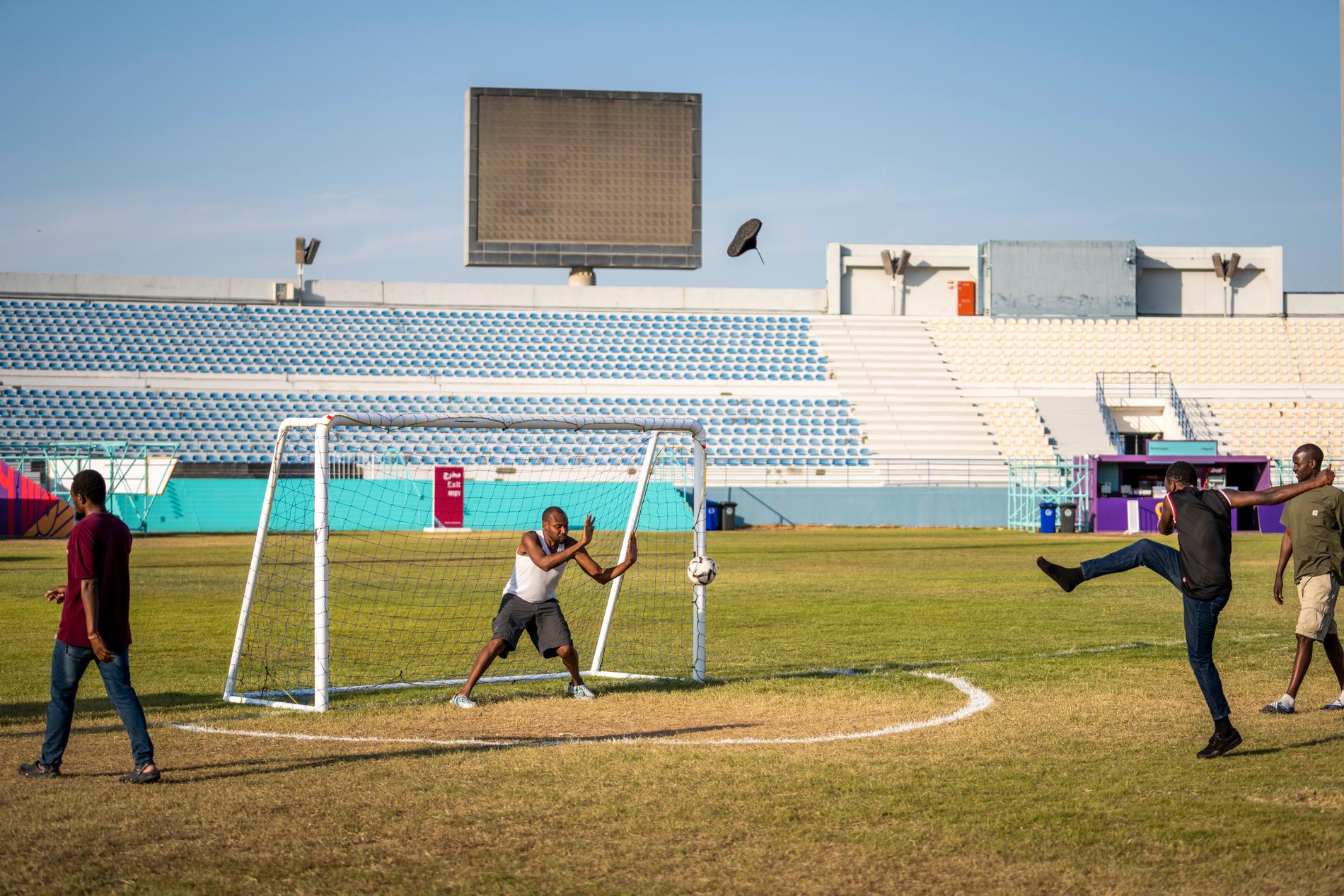 Migrantarbetare samlas vid Asian Cricket stadium i Asian town strax vänster om Doha för att på storbildskärm följa matcher under Fotbolls-VM i Qatar 2022. Mellan matcherna spelar de fotboll på en liten plan. 