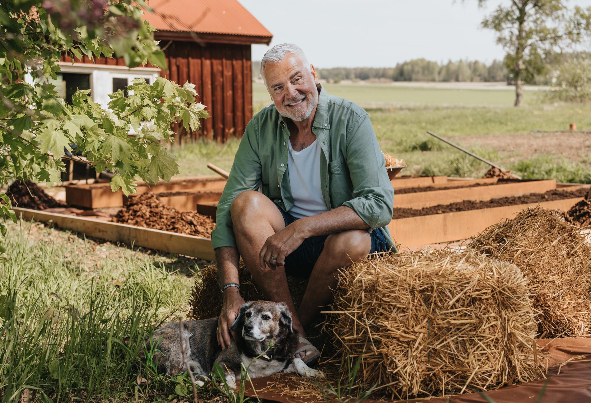 Ernst Kirchsteiger sitter bland höbalar, gräs och jord och klappar en hund