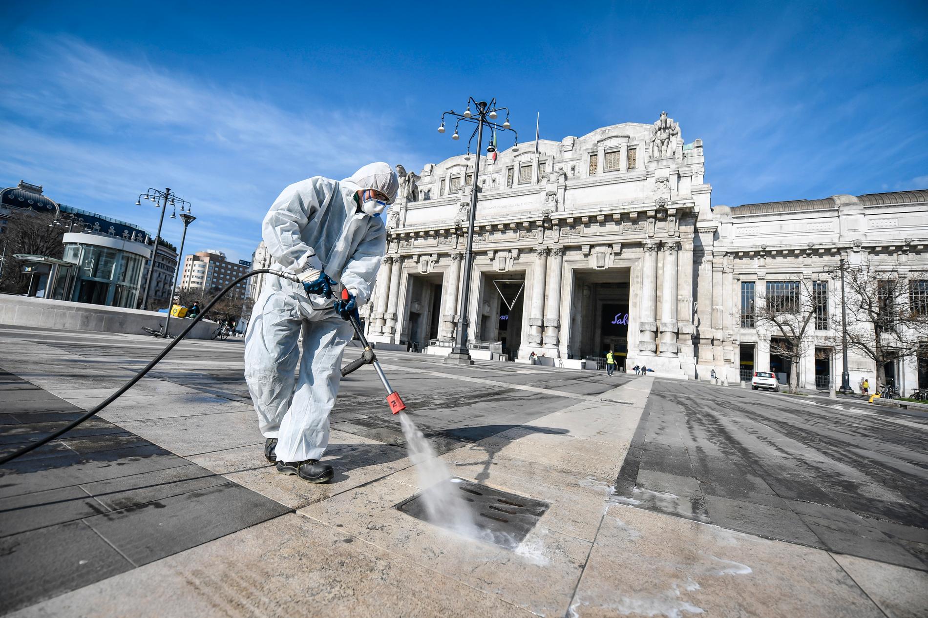 En skyddsklädd arbetare desinficerar ett område framför järnvägsstationen i Milano i Italien – en plats som drabbats hårt av det nya coronaviruset.