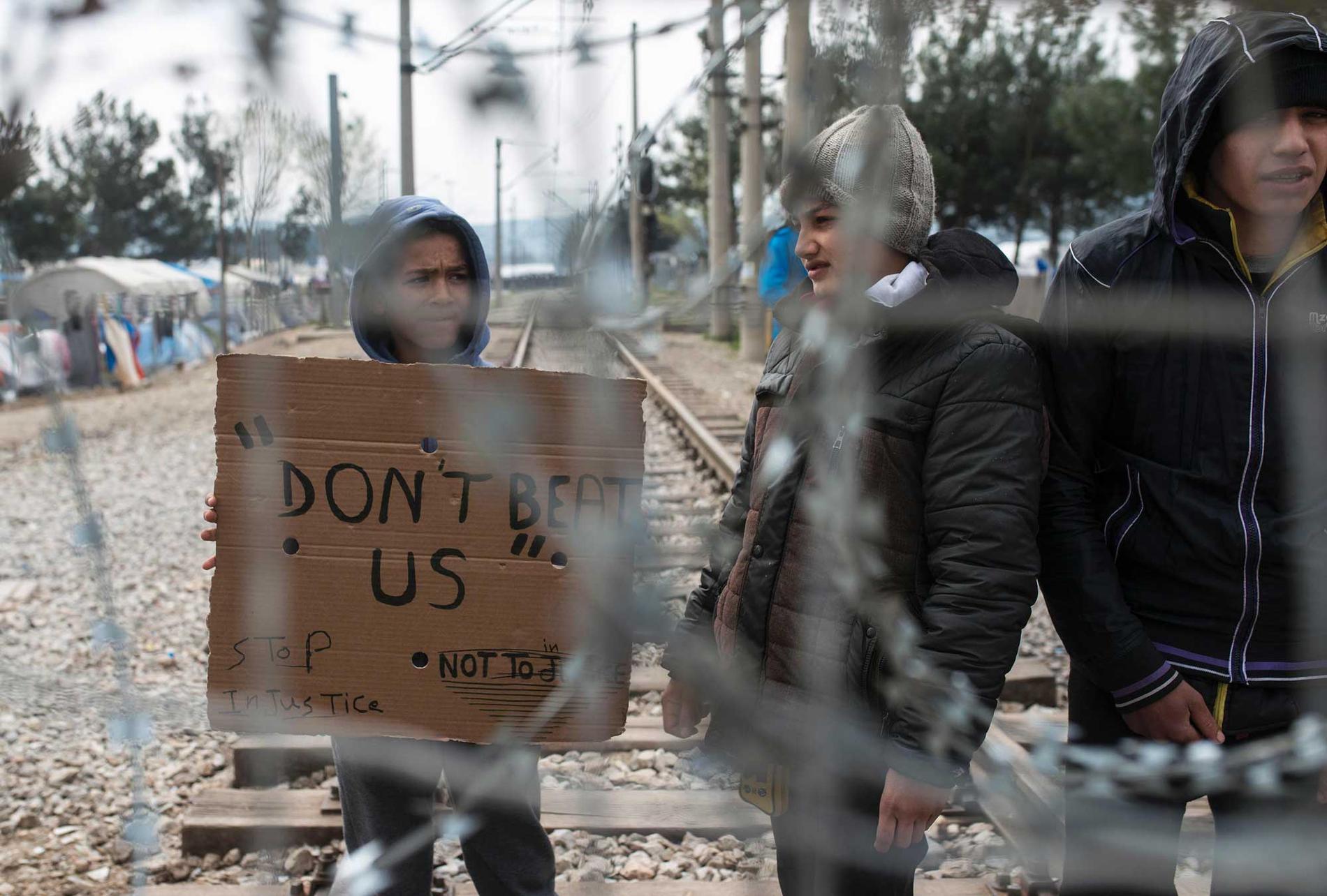 Hundratals flyktingar protesterade under söndagen mot stängda gränser vid gränsstaketet mellan Grekland och Makedonien. Här håller en pojke upp ett plakat med texten ”slå oss inte”. Samtidigt motades andra personer av polis.