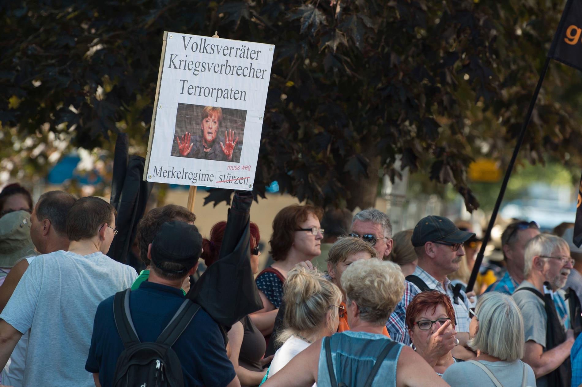 Demonstranter håller upp plakat med ett foto av Angela Merkel och texten "Förrädare, krigsförbrytare terroristgudmor – Merkelregeringen måste bort" i samband med Merkels besök i Dresden.