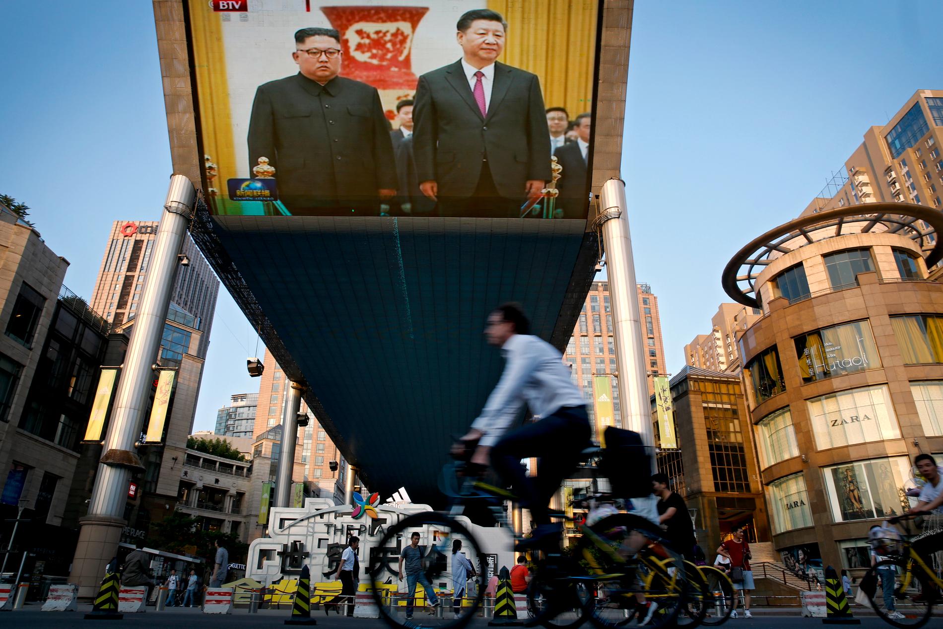 Cyklister passerar en stor tv-skärm i Peking med en sändning från ceremonin där Nordkoreas ledare Kim Jong-Un välkomnades av Kinas president Xi Jinping i Folkets stora sal på tisdagen.