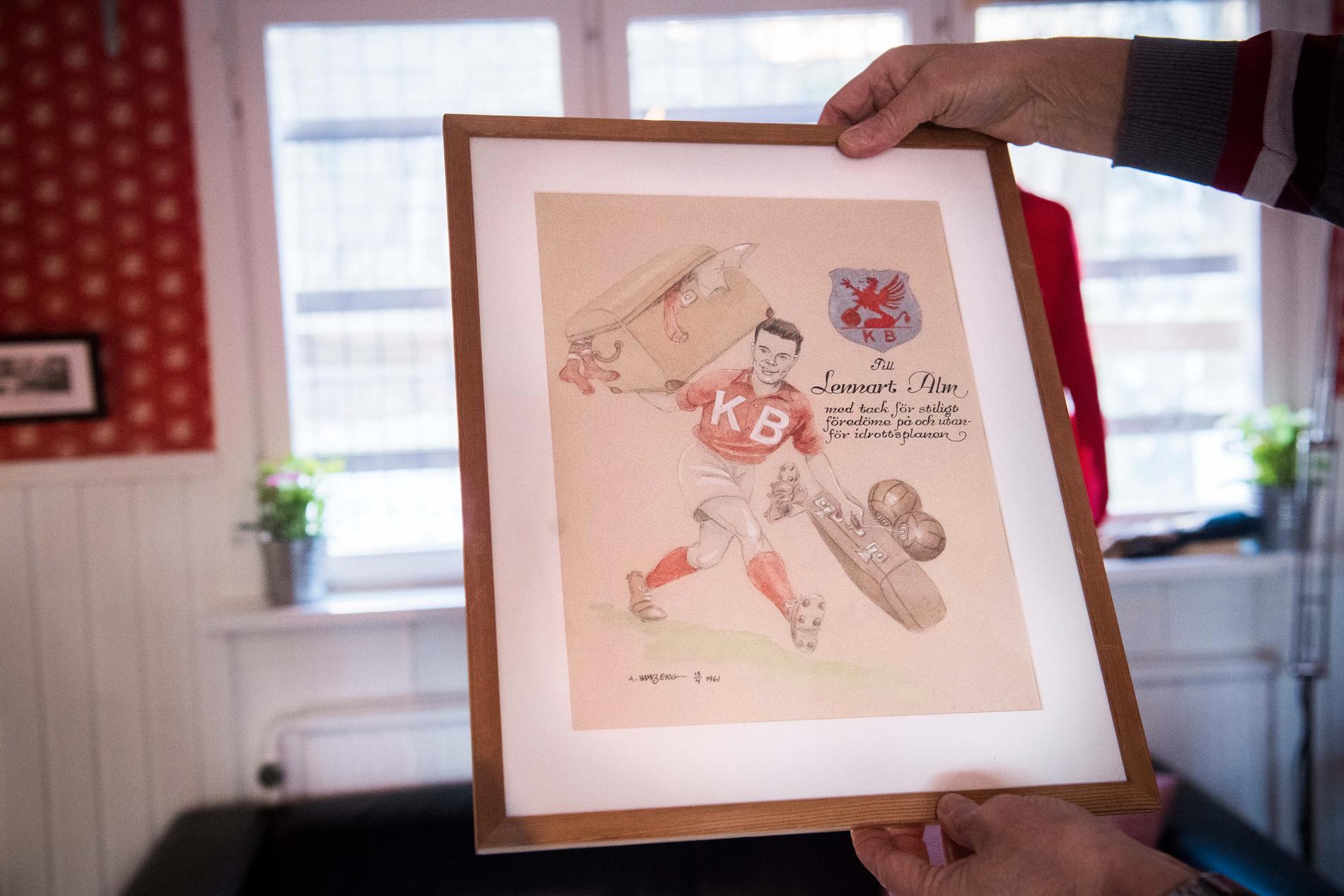 Tavlan föreställandes Lennart Alm i KB:s rödvita fotbollsställ – glatt kånkandes på ett par materialväskor – tecknades av klubbens grundare ”Acke” Hamberg år 1961.