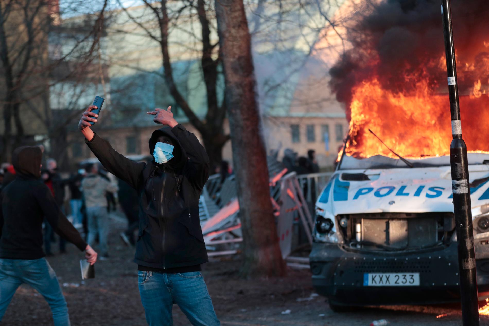 En person  tar en selfie samtidigt som  han visar långfingret framför en brinnande polisbil i Örebro på långfredagen. 