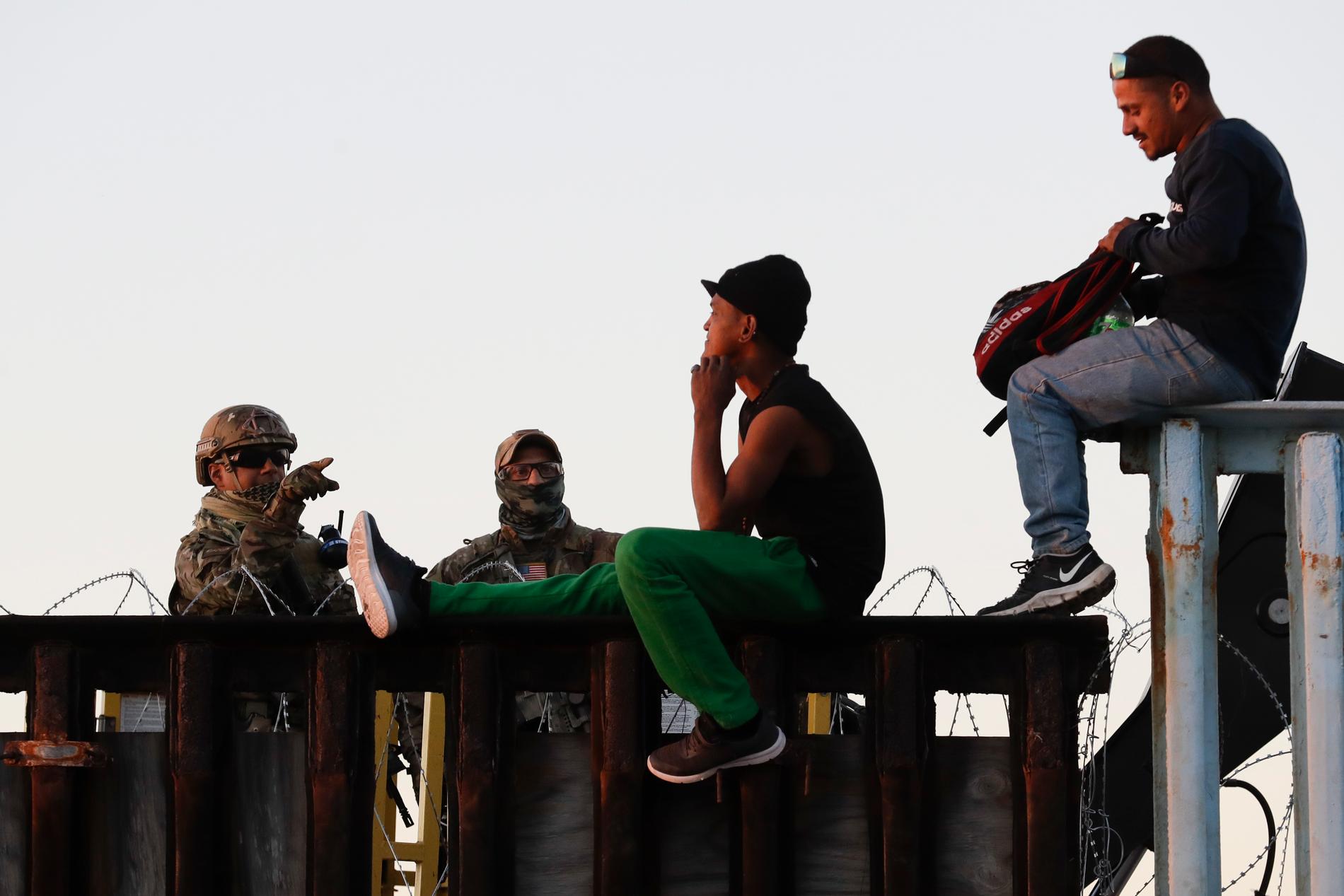 Amerikanska gränspoliser i samspråk med två migranter som sitter uppe på gränsstaketet, på andra sidan taggtråden.