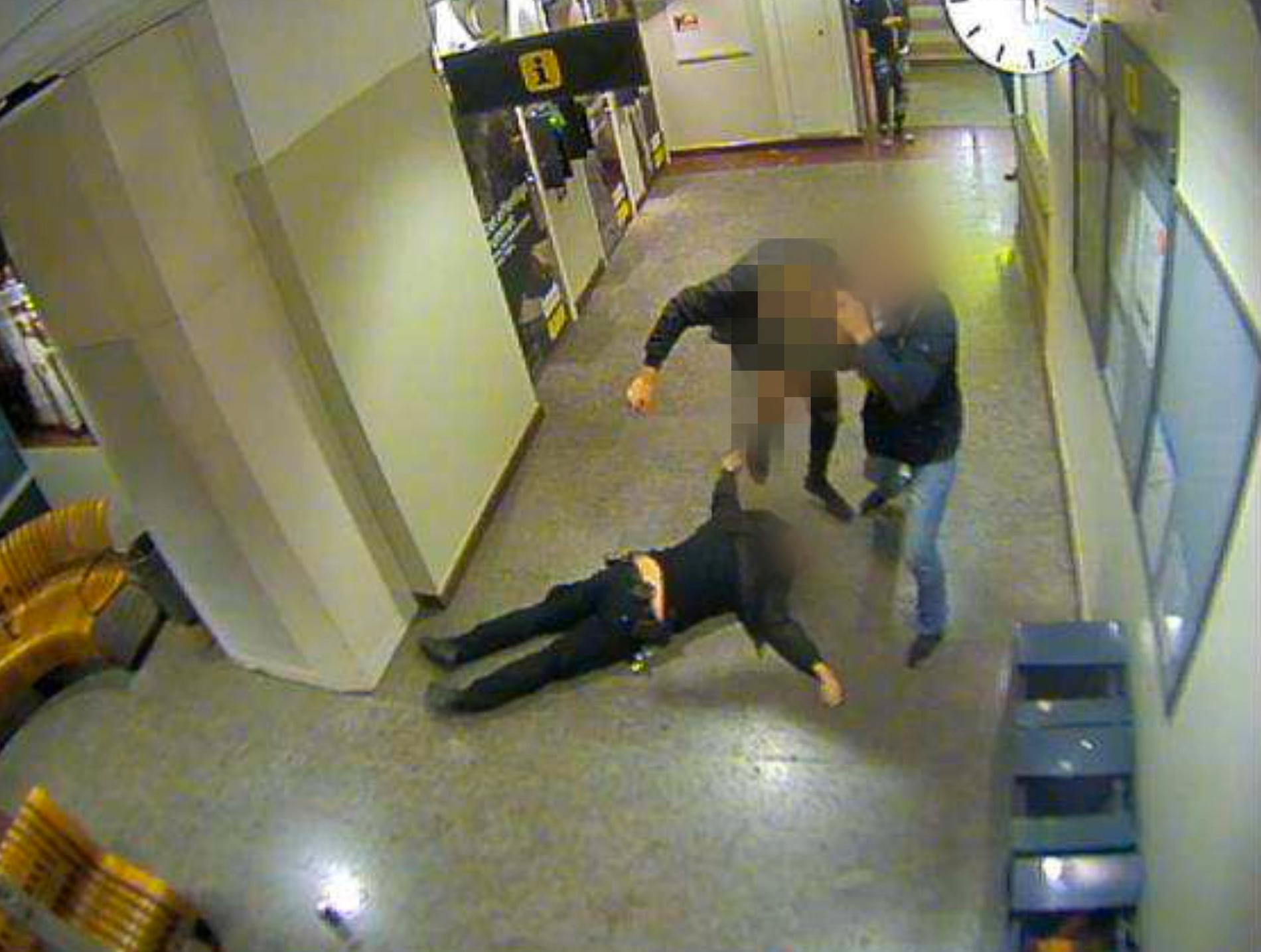 Mannen ses lyfta sitt ben för att sparka polismannen i huvudet samtidigt som en annan man ur gruppen försöker dra bort honom. 