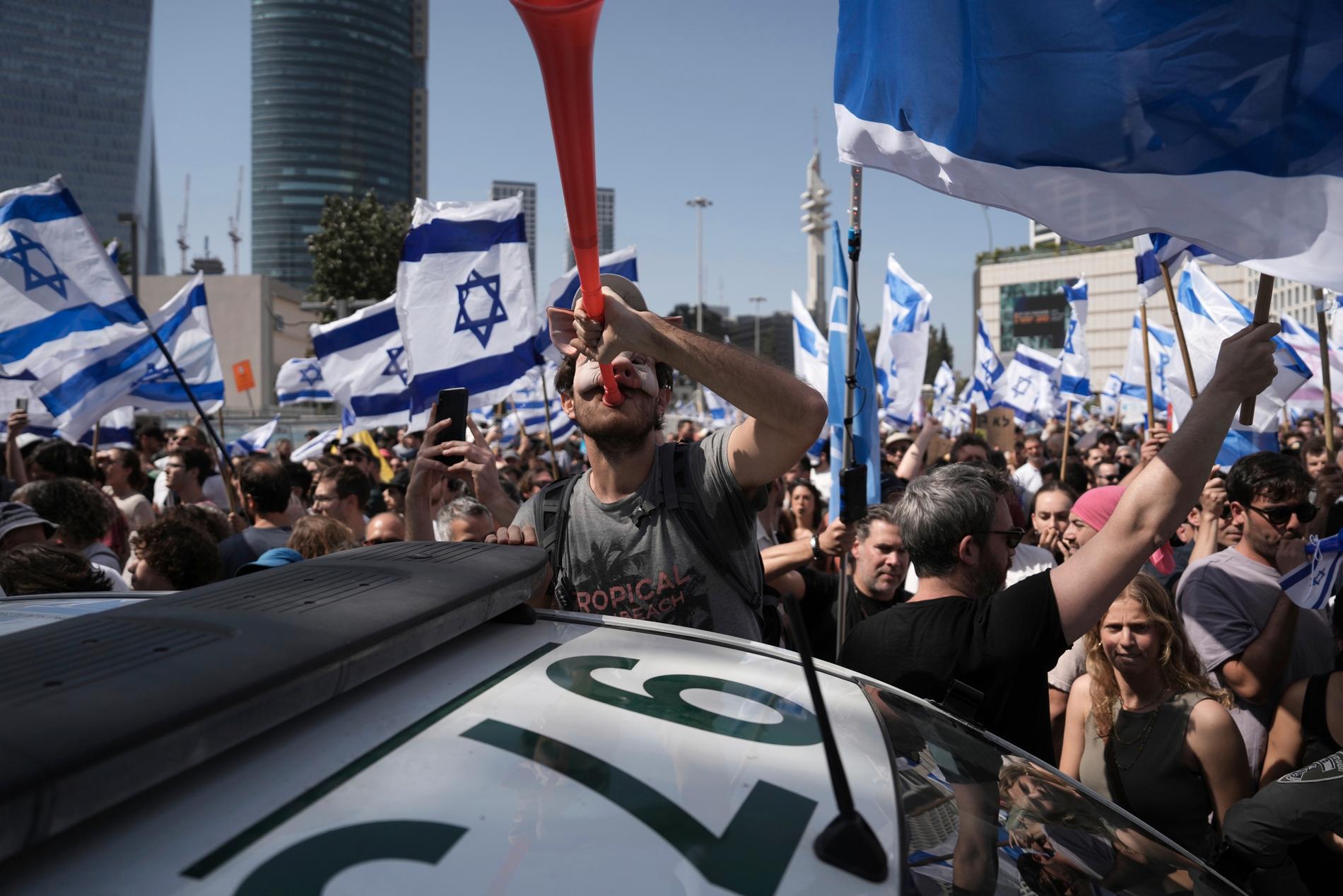 En demonstrant i grismask blåser i en vuvuzela bredvid en polisbil i Tel Aviv.