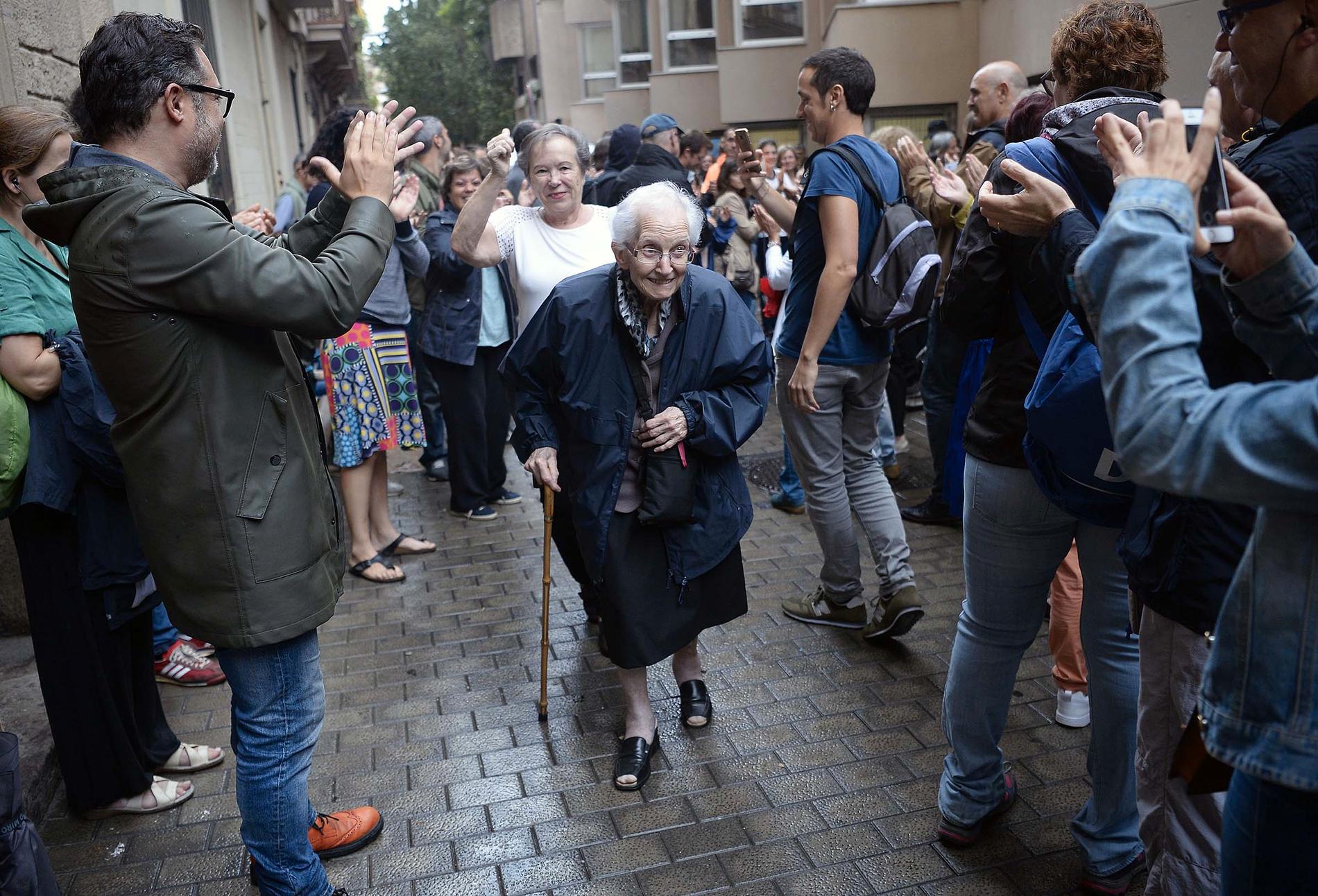 Människor vid en vallokal applåderar en äldre kvinna som varit och röstat.