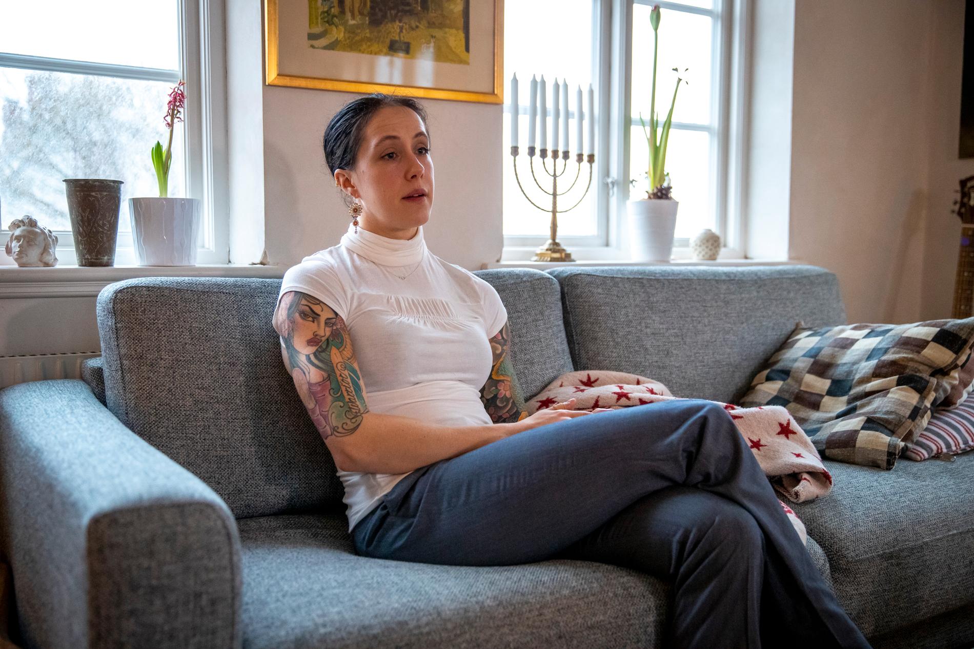 Någonstans i Sverige sitter en ung kvinna i ljus blus och gråa byxor på en soffa.