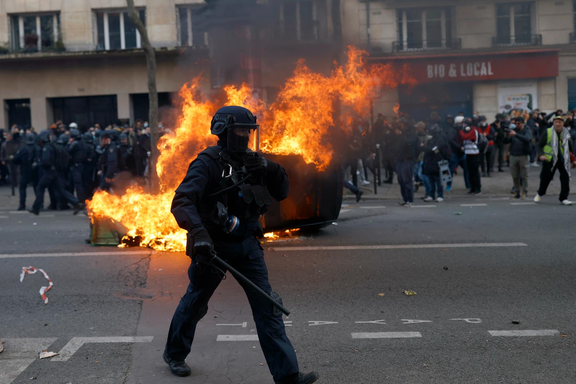 En kravallpolis går förbi en brinnande bil under sammandrabbningar vid en demonstration.