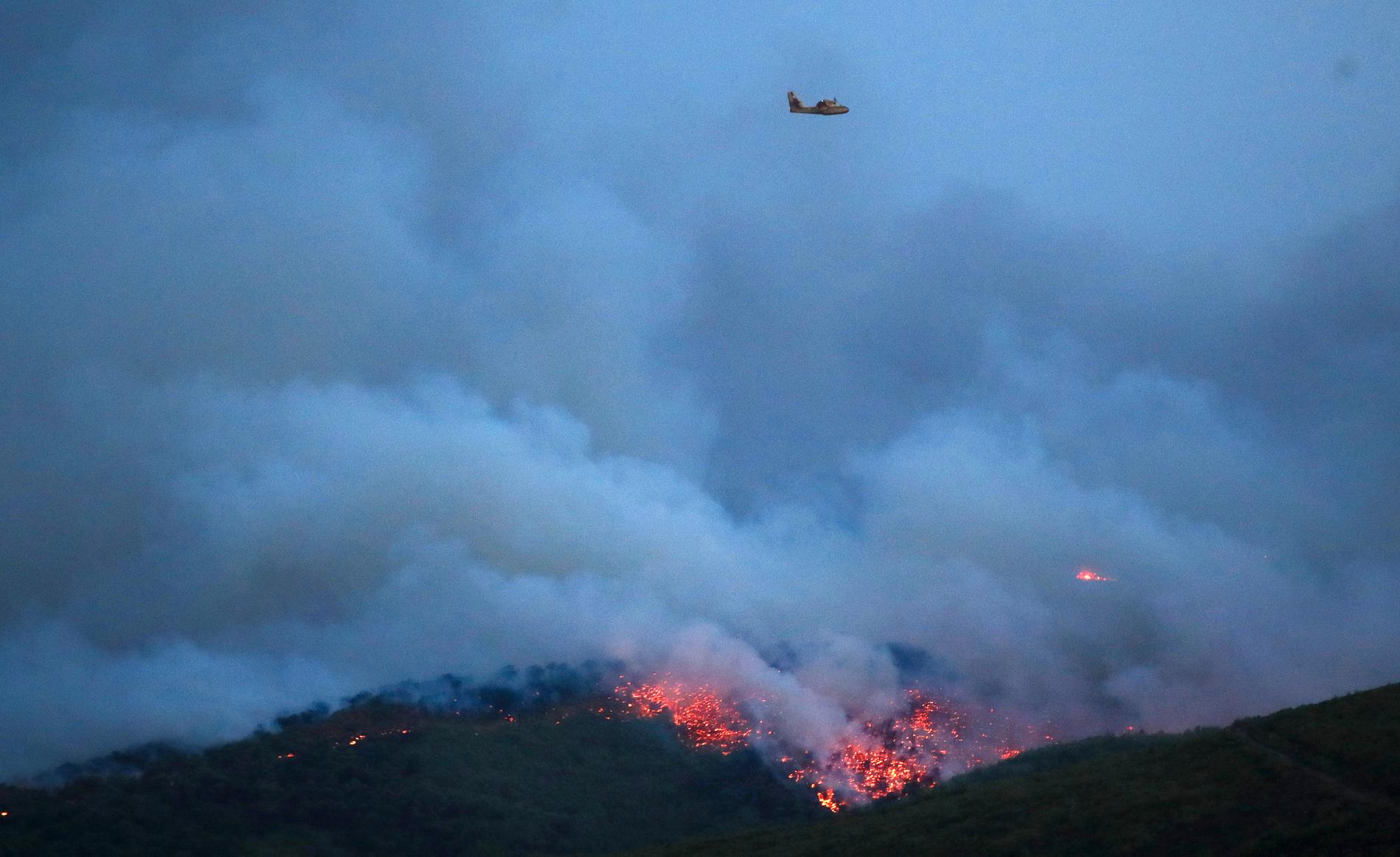 Ett flygplan flyger över röken och elden som kommer från staden Mati, öster om Aten.