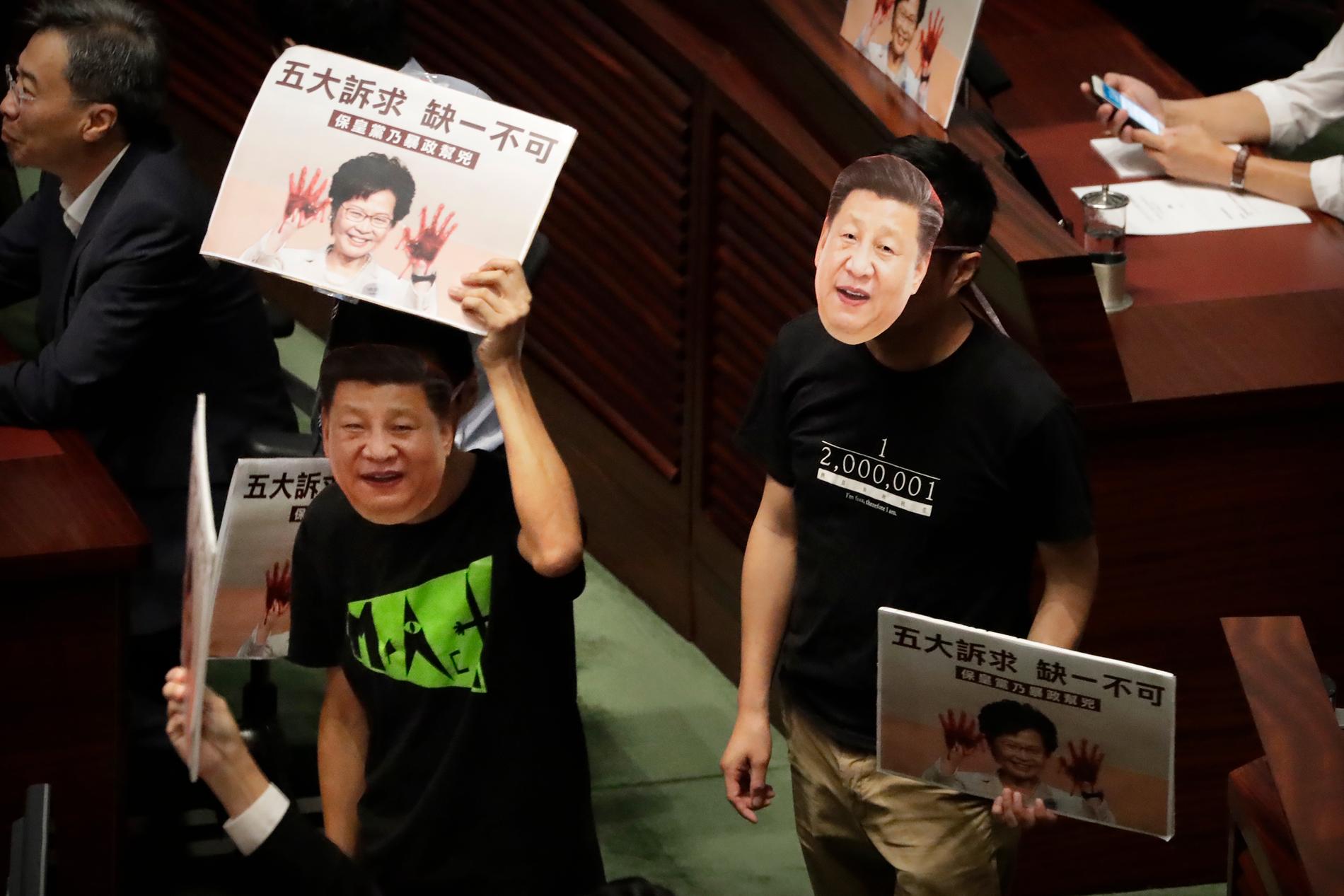 Demokrativänliga parlamentsledamöter bar masker föreställande Kinas president Xi Jinping när de avbröt Carrie Lams tal.