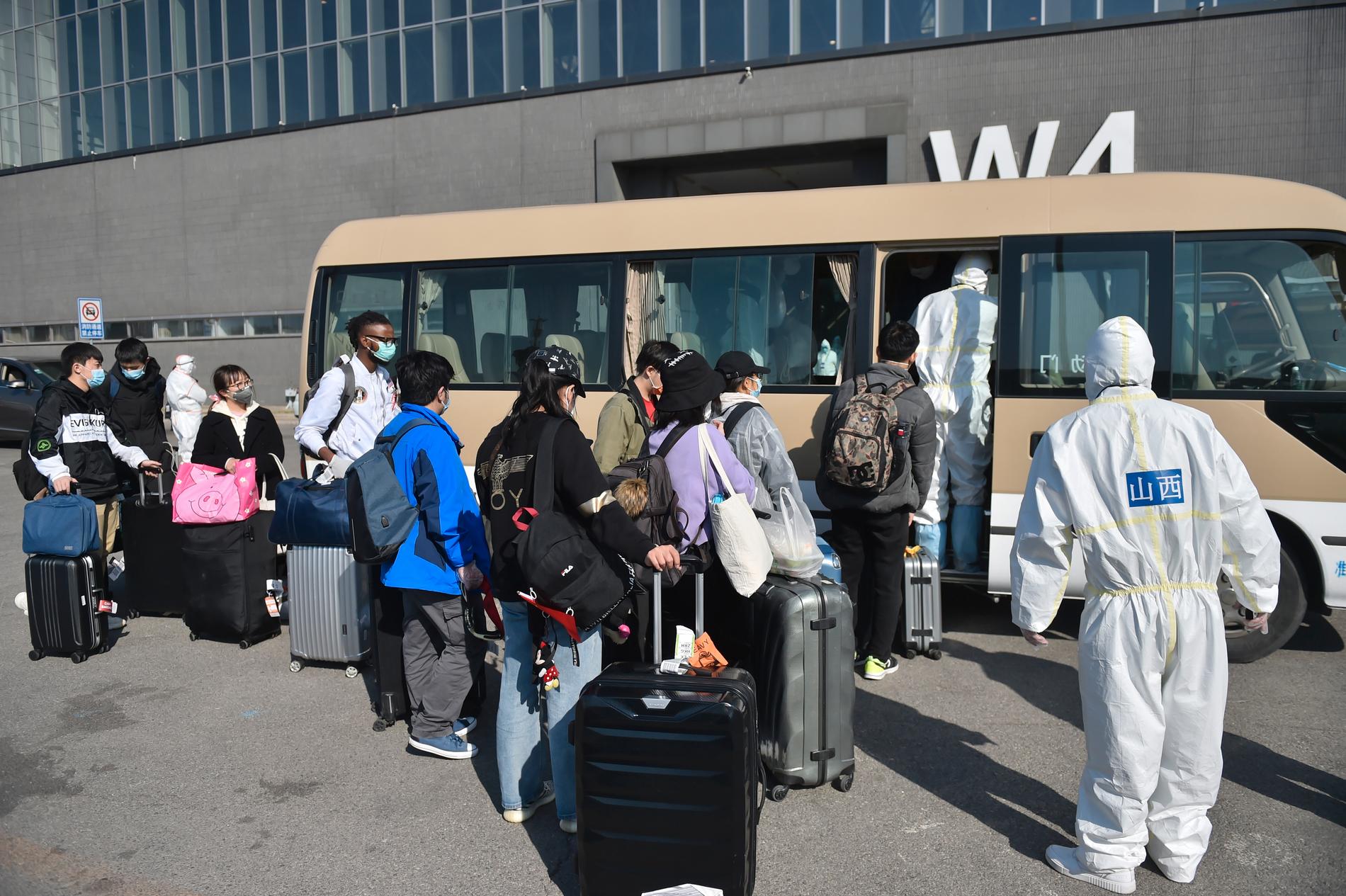 Smittskyddsarbetare ser på när resenärer kliver på en buss vid en utställningshall som gjorts om till en anläggning för att kontrollera personer som anlänt med flyg till Peking.