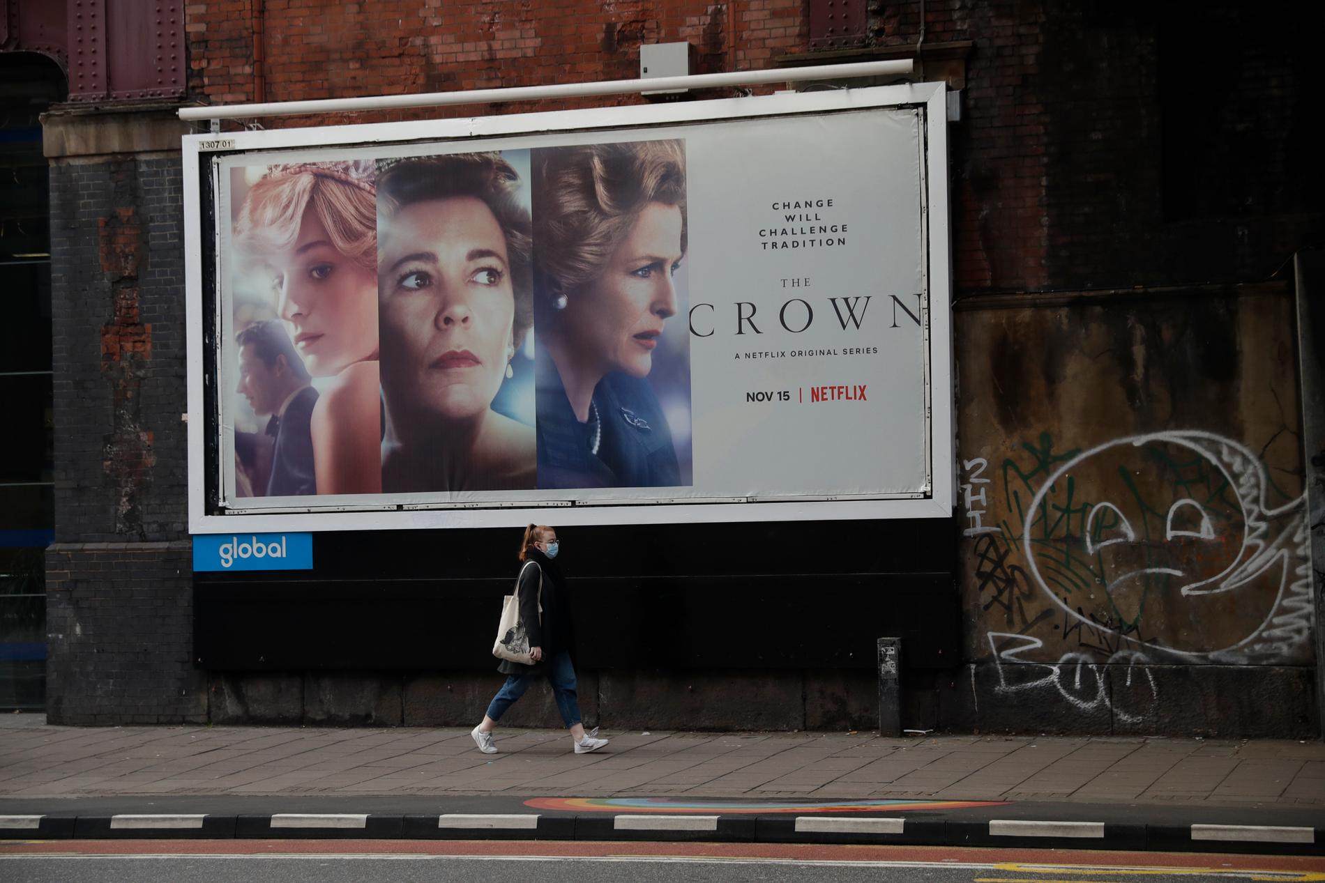 En kvinna i ansiktsmask går förbi en stor reklamskylt för serien ”The Crown” i London.