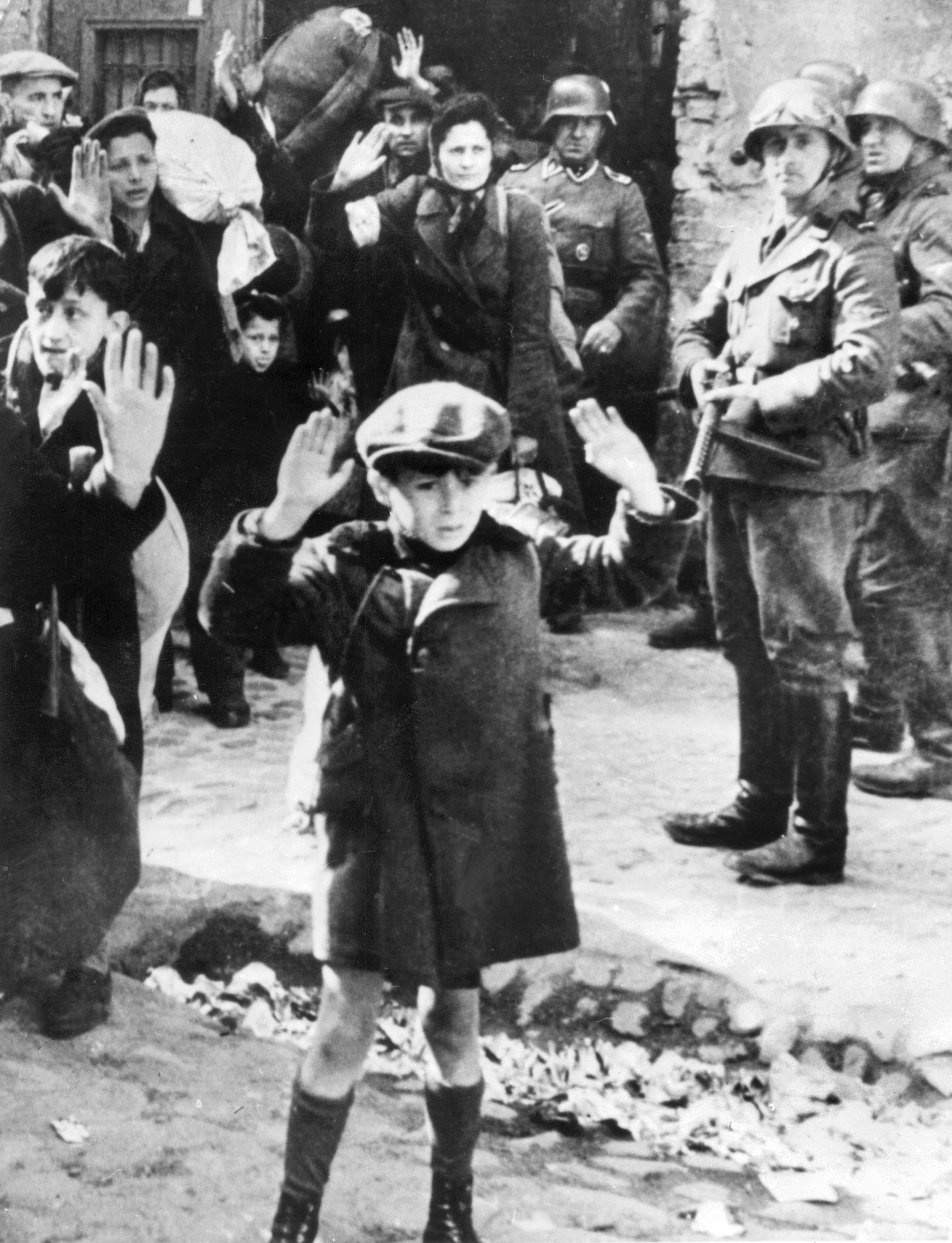 En liten pojke bland andra barn och vuxna som håller upp händerna när de omringas av tyska soldater med gevär i gettot i Warszawa under andra världskriget.