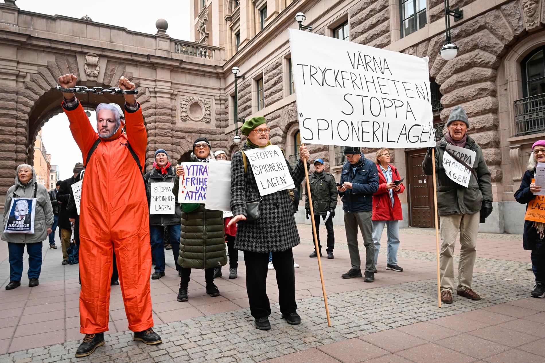 Utanför riksdagen demonstreras det i dag mot grundlagsändringen. En demonstrant har en mask föreställande Julian Assange. På plakaten står bland annat ”Värna tryckfriheten – stoppa spionerilagen”.