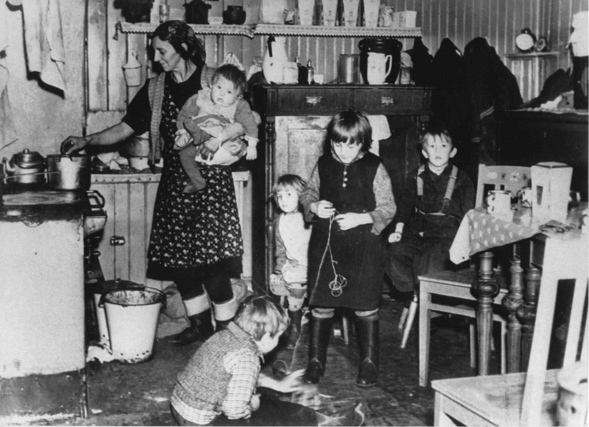Fattig-Sverige. Arbetarfamilj i Stockholm på 40-talet. Kvinna i förkläde och med barn på armen står vid spisen och lagar mat . Fyra barn leker på köksgolvet.