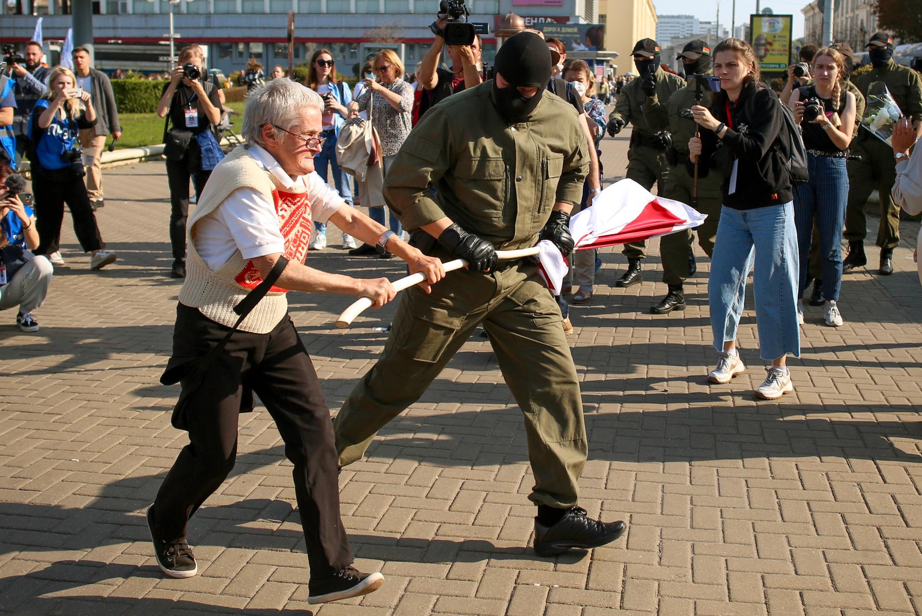 En polis försöker rycka den vit-röd-vita oppositionsflaggan ur 73-åriga Nina Bahinskaja händer underprotester i Minsk i Belarus på lördagen. Bahinskaja uppges sedan ha gripits.