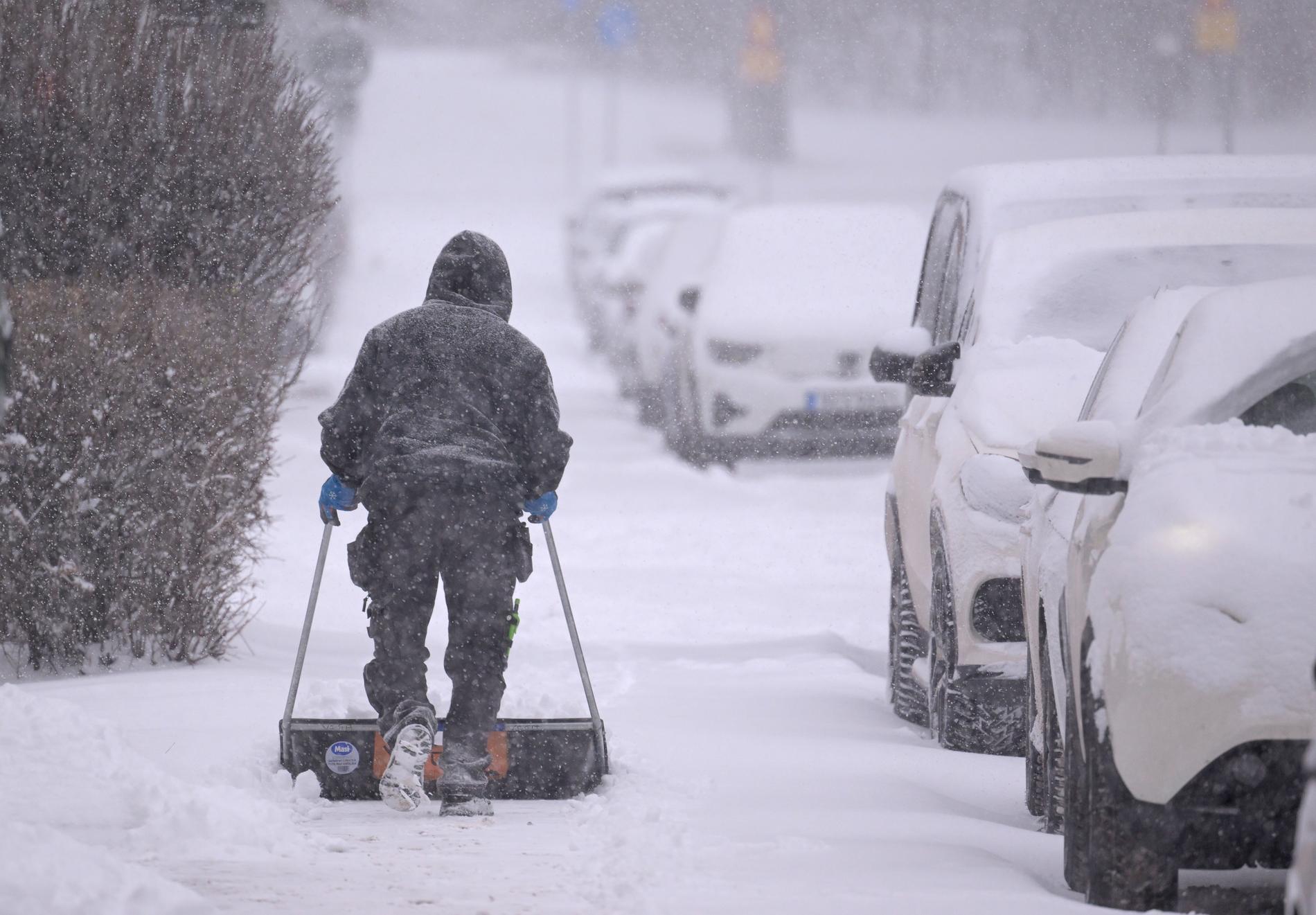 En man tar bort snö med en snöskovel på trottoaren mellan ett bostadshus och snötäckta parkerade bilar. Bild från november.