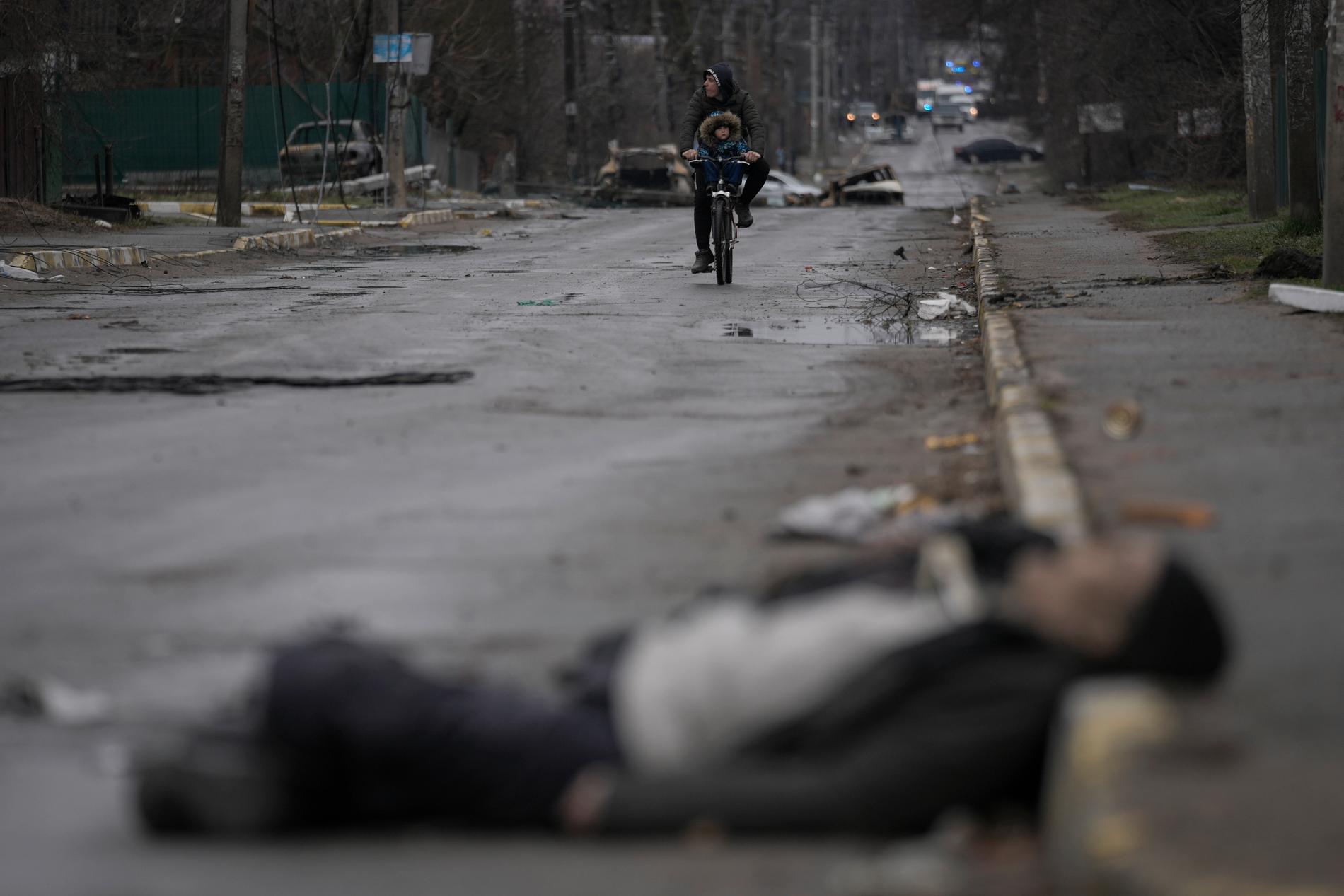 En man och ett barn på en cykel på en gata där en död person lämnats kvar efter de ryska styrkorna. 